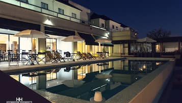 Zona de piscina de hotel con sillones reclinables y sombrillas, piscina cristalina y un edificio con varios pisos y balcones.