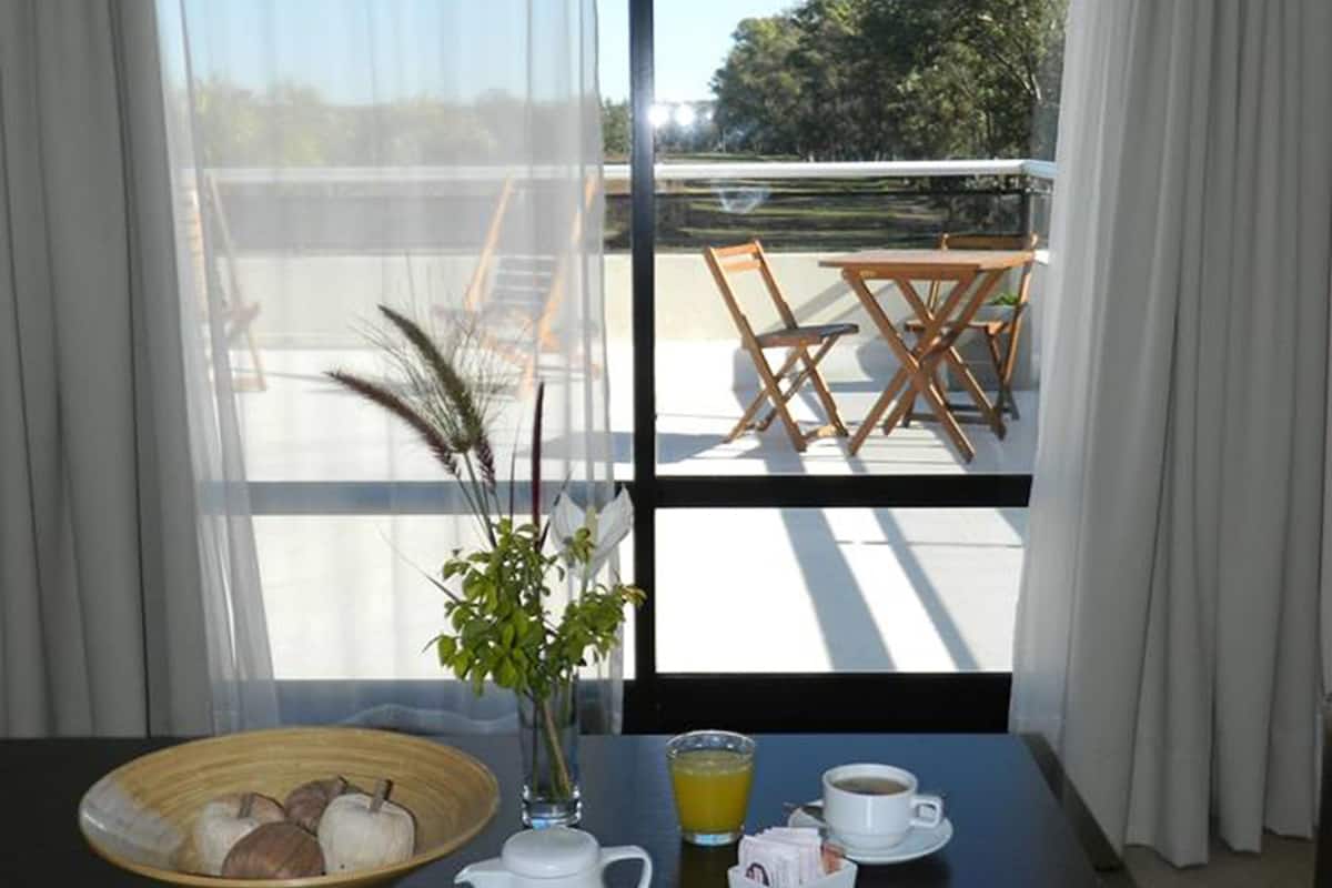 A breakfast setup with a bowl of fruit, a vase with a plant, a pitcher, a glass of orange juice, and cups of coffee and tea on a table by a window with a view of a balcony and outdoor furniture.