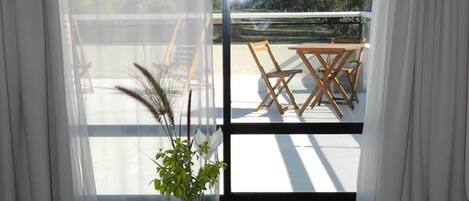 Una preparación para el desayuno con un bol de fruta, un florero con una planta, una jarra, un vaso de jugo de naranja y tazas de café y té sobre una mesa junto a una ventana con vistas a un balcón y mobiliario exterior.