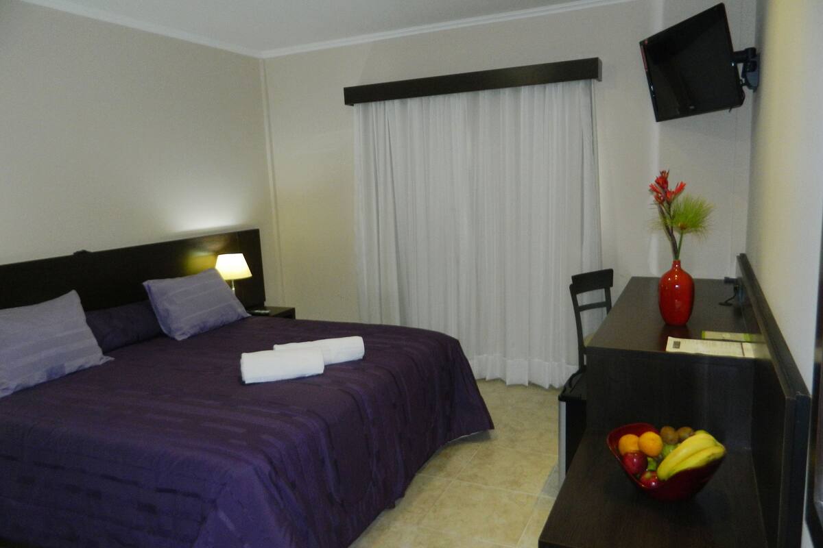 A hotel room with a bed, a desk with a vase and fruit bowl, a TV mounted on the wall, and a window with curtains.