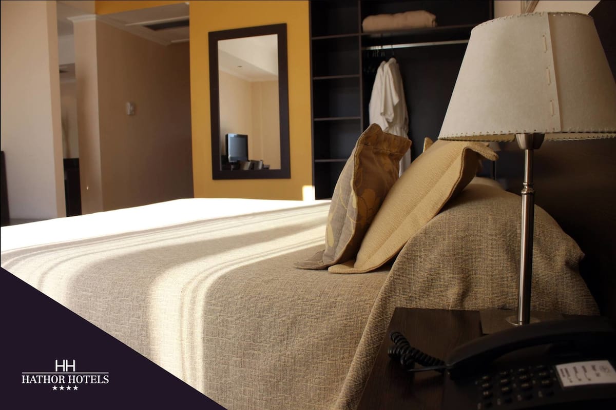 A hotel room with a neatly made bed, a bedside lamp, a telephone, and a mirror on the wall.