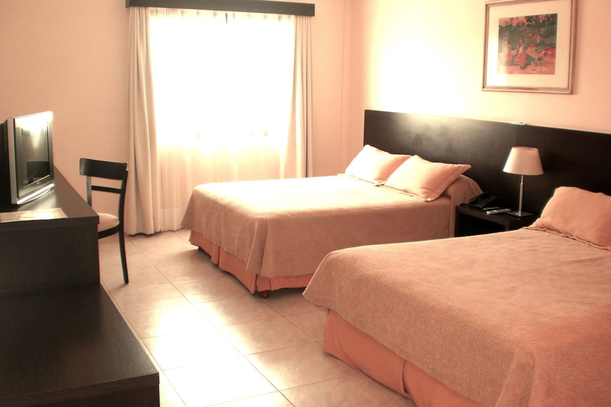 A hotel room with two beds, a desk with a television, a chair, a nightstand with a lamp, and a painting on the wall.