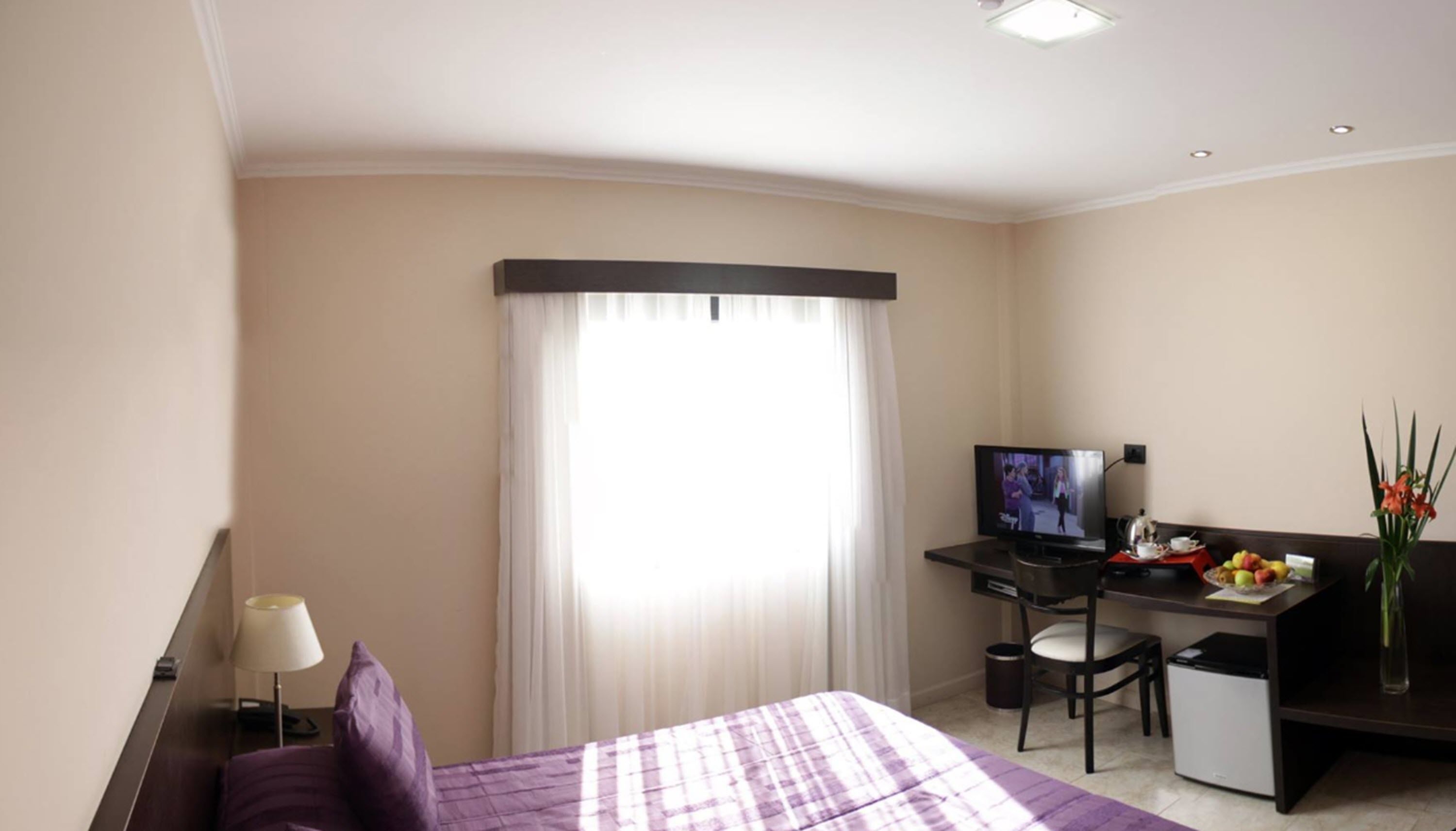 Ein Hotelzimmer mit einem Bett, einem Schreibtisch mit Computer, einem Fernseher, einer kleinen Mülltonne und einem Fenster mit Vorhängen.