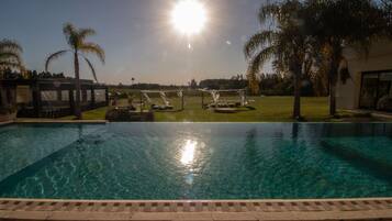 A clear swimming pool with a sunlit sky, palm trees, and a grassy area with a gazebo.