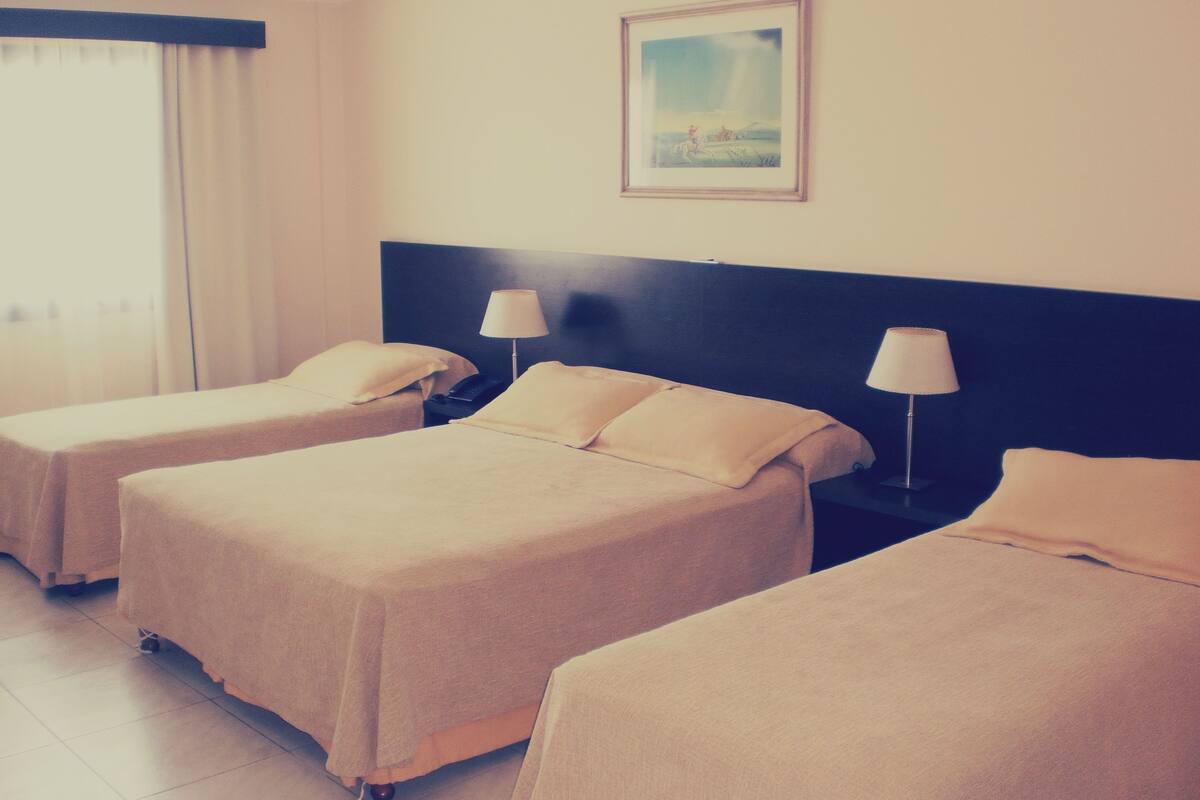 A hotel room with two single beds, a nightstand with a lamp, a painting on the wall, and a window with curtains.