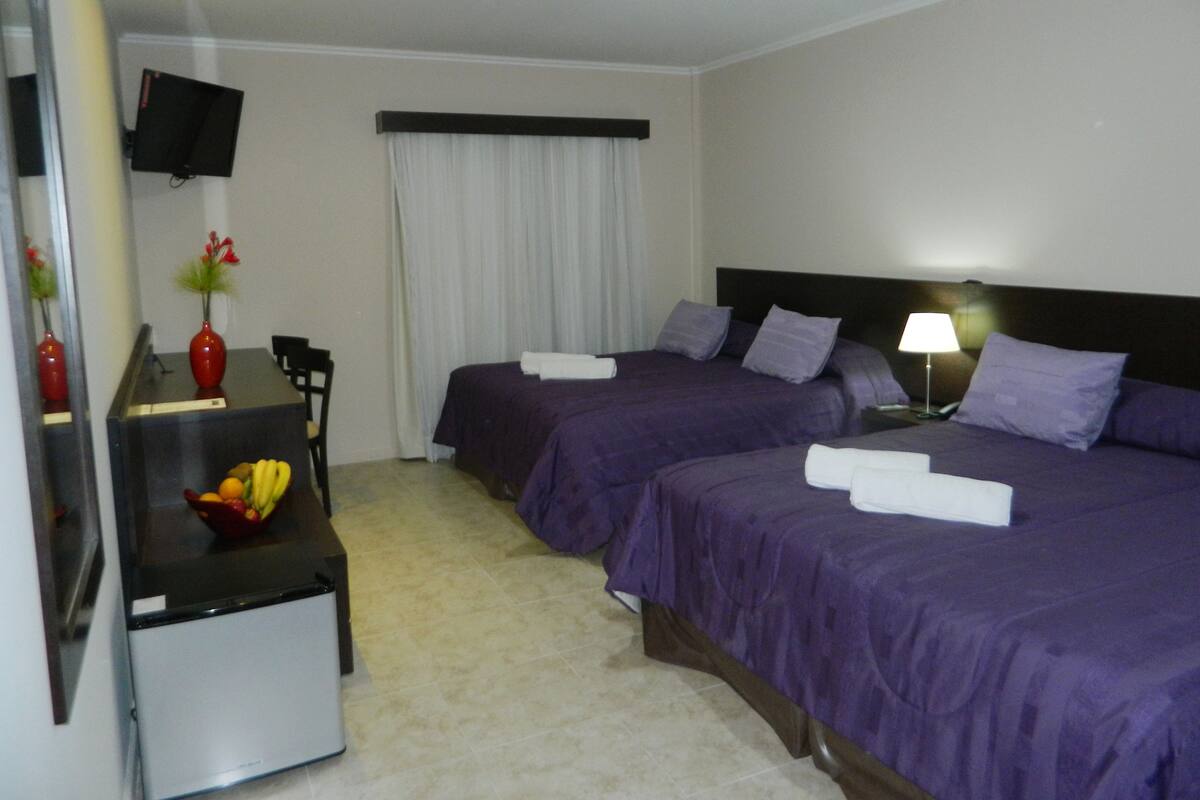 A hotel room with two beds, a desk with a chair, a TV, and a fruit bowl.