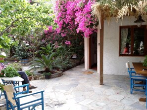 A patio with stone flooring, blue chairs, and tables. There are potted plants and a hanging basket with pink flowers. - Olive Tree Cottages (Kantanos-Selino)