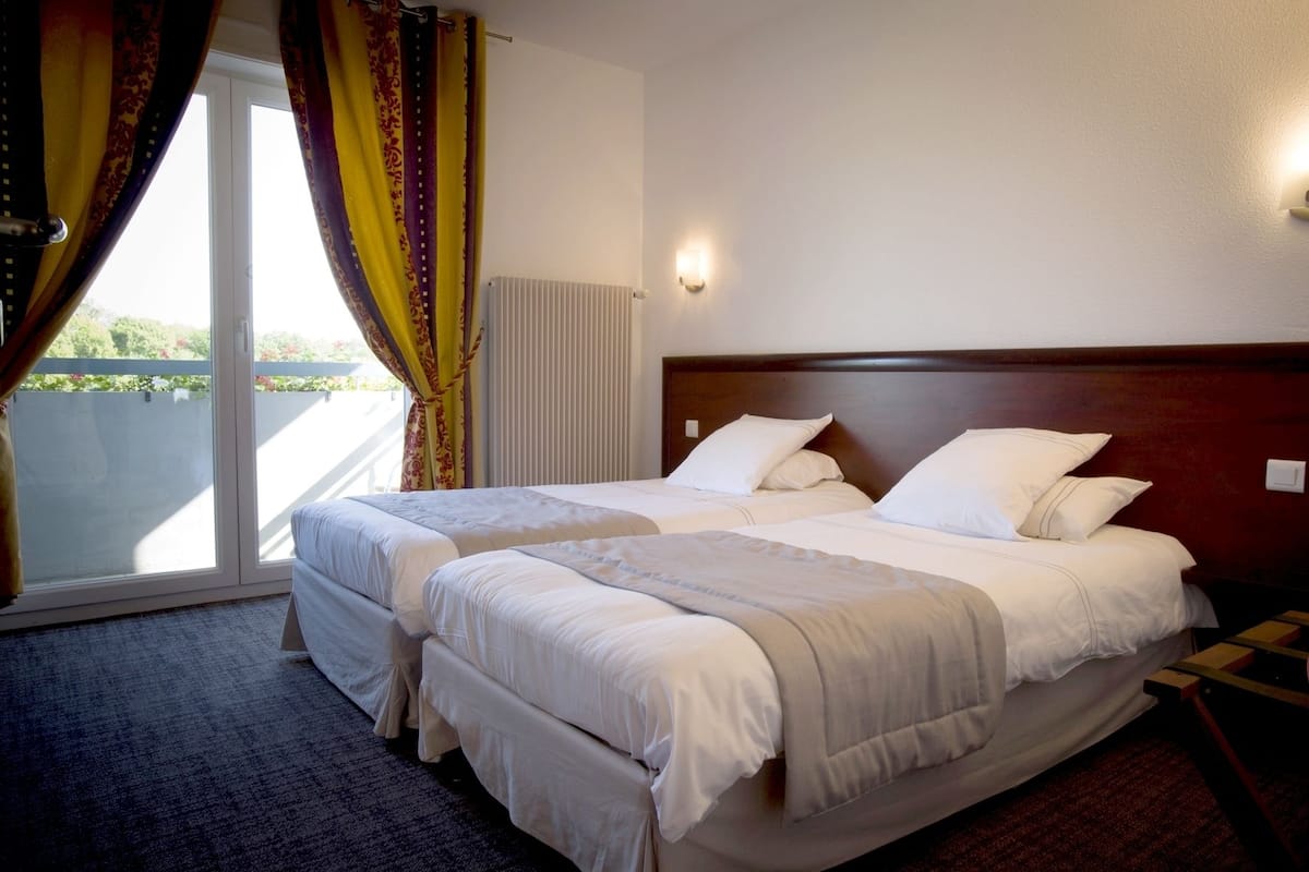 A hotel room with two beds, a large window with curtains, a radiator, and a wall-mounted light.
