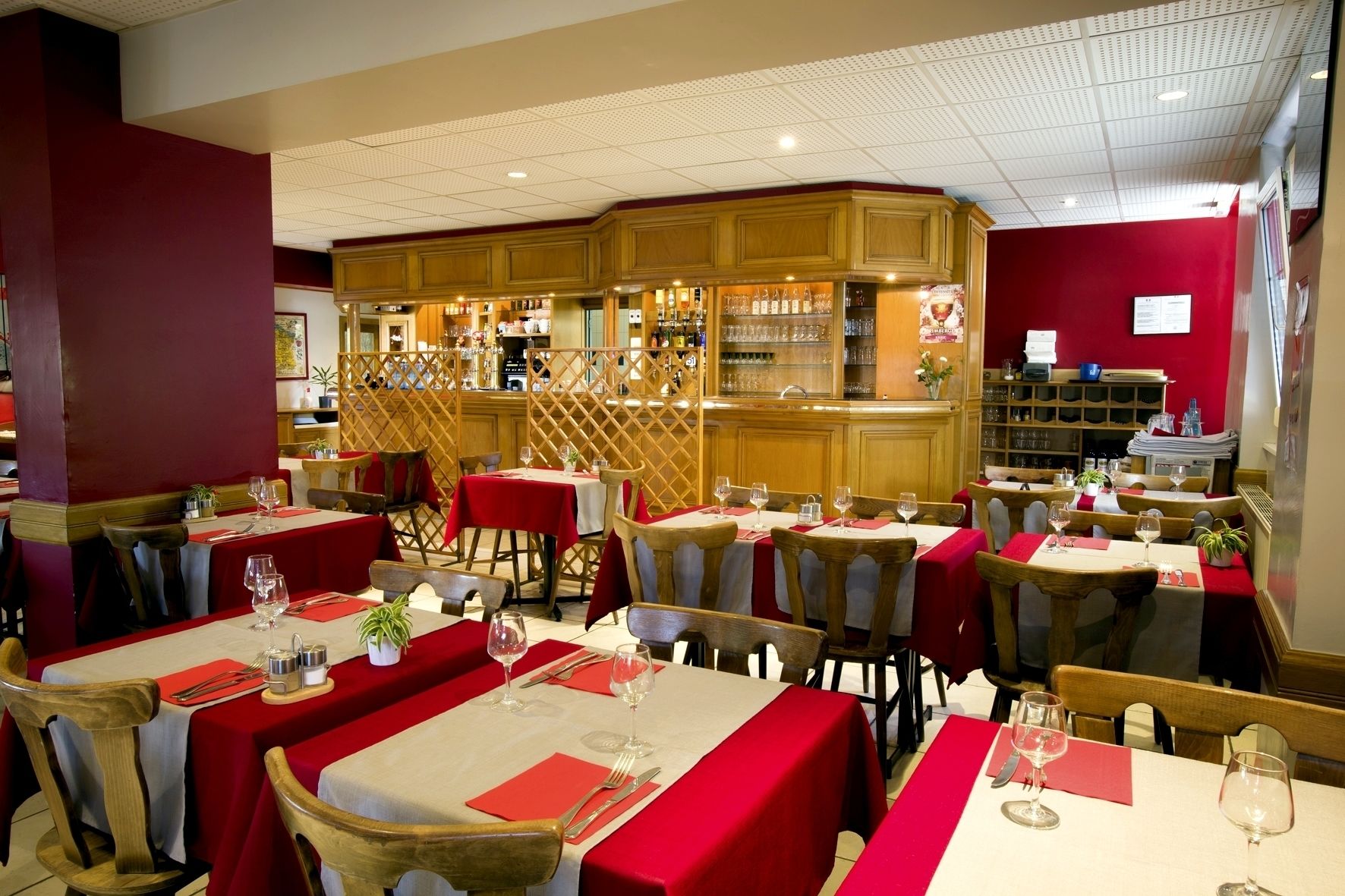 A restaurant with multiple tables set with red tablecloths and white table settings, wooden chairs, and a bar area with shelves of glasses and bottles.