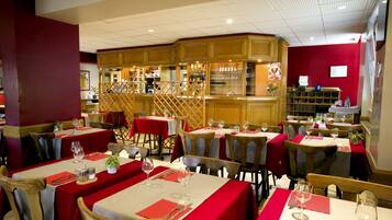 A restaurant with multiple tables set with red tablecloths and white table settings, wooden chairs, and a bar area with shelves of glasses and bottles.