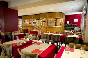 A restaurant with multiple tables set with red tablecloths and white table settings, wooden chairs, and a bar area with shelves of glasses and bottles.