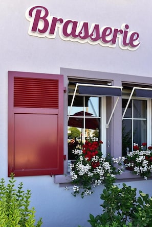 A building with a sign reading 'Brasserie' above a window with red shutters and potted plants with red and white flowers.