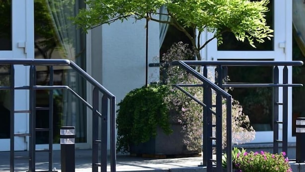 A building with a staircase leading to a door, a small tree, and a flower bed. - Au Lion d'Or (Burnhaupt-le-Haut)