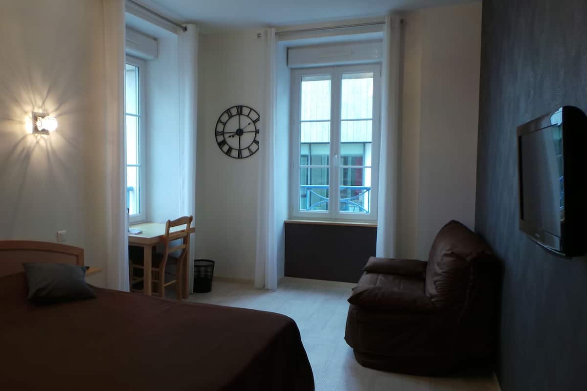 A bedroom with a bed, a chair, a desk, a clock, and a TV.