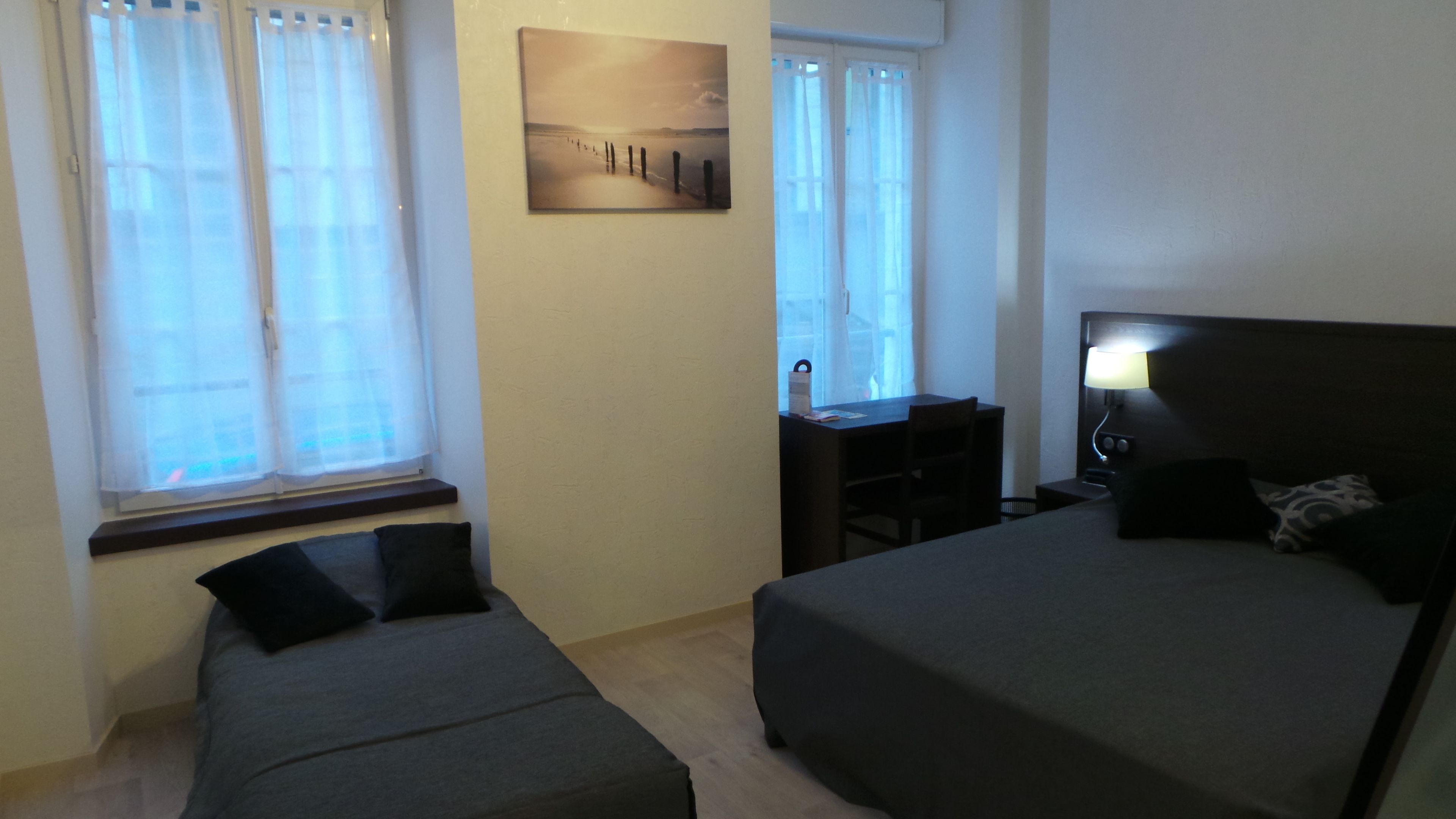 Quarto de hotel com cama, escrivaninha e cadeira. Há uma janela com cortinas, um quadro na parede e uma luminária na mesa de cabeceira.