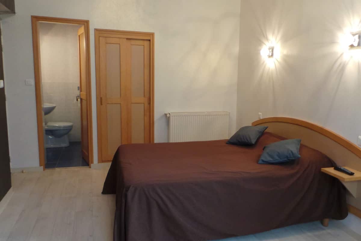 A bedroom with a bed, a nightstand, a wall-mounted light, and a door leading to a bathroom.