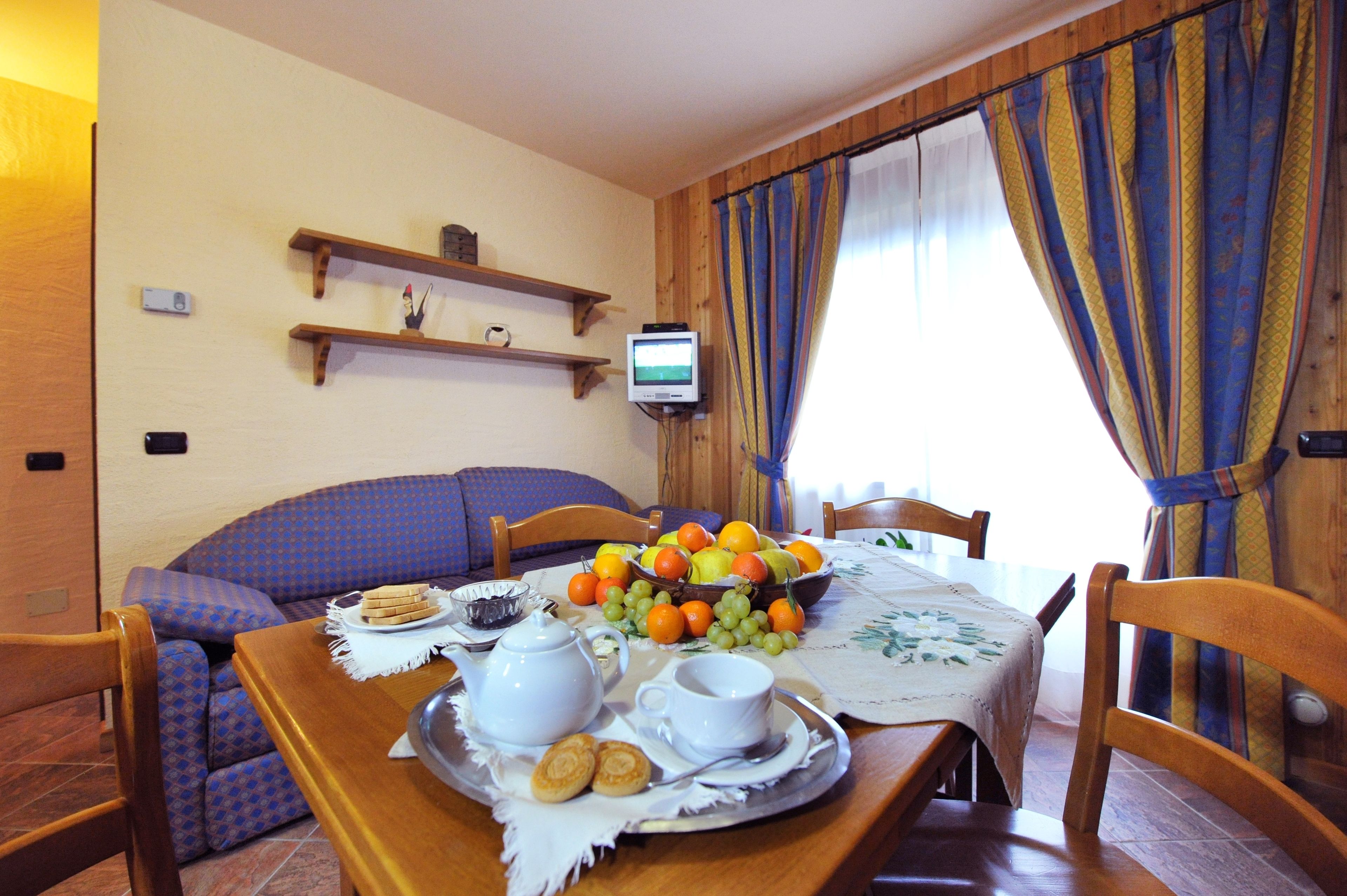 Comedor con una mesa dispuesta para el desayuno, que incluye un bol de fruta, un hervidor de agua, tazas y galletas. Hay sillas de madera, un sofá y un televisor sobre una estantería.