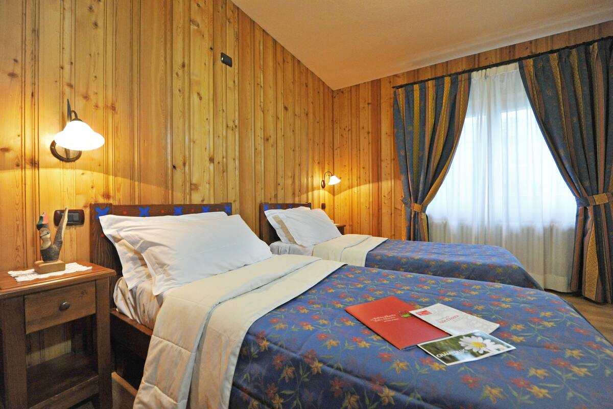 A hotel room with wooden panel walls, two beds with floral bedding, bedside lamps, a nightstand with a phone, and a window with curtains.