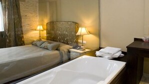 Habitación de hotel con una cama individual, una bañera grande, dos lámparas de noche, un mesita de noche de madera y un espejo.