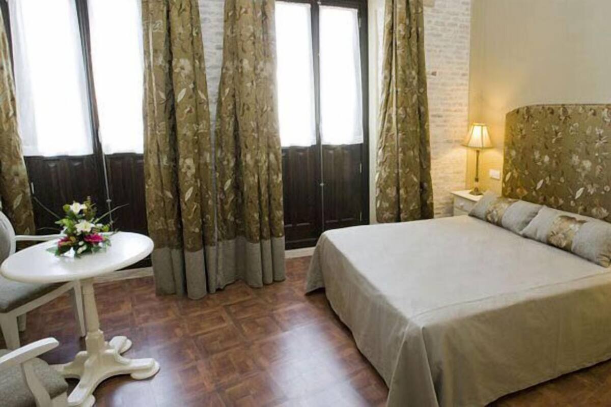 A hotel room with a bed, a small table with a flower arrangement, a nightstand with a lamp, and curtains.