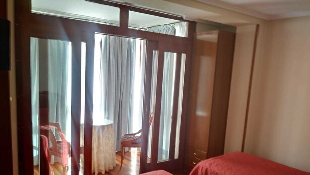 A room with two beds, a wooden wardrobe, a small table with a chair, and a window with curtains. - Hostal Sancho Garcia (Espinosa de los Monteros)
