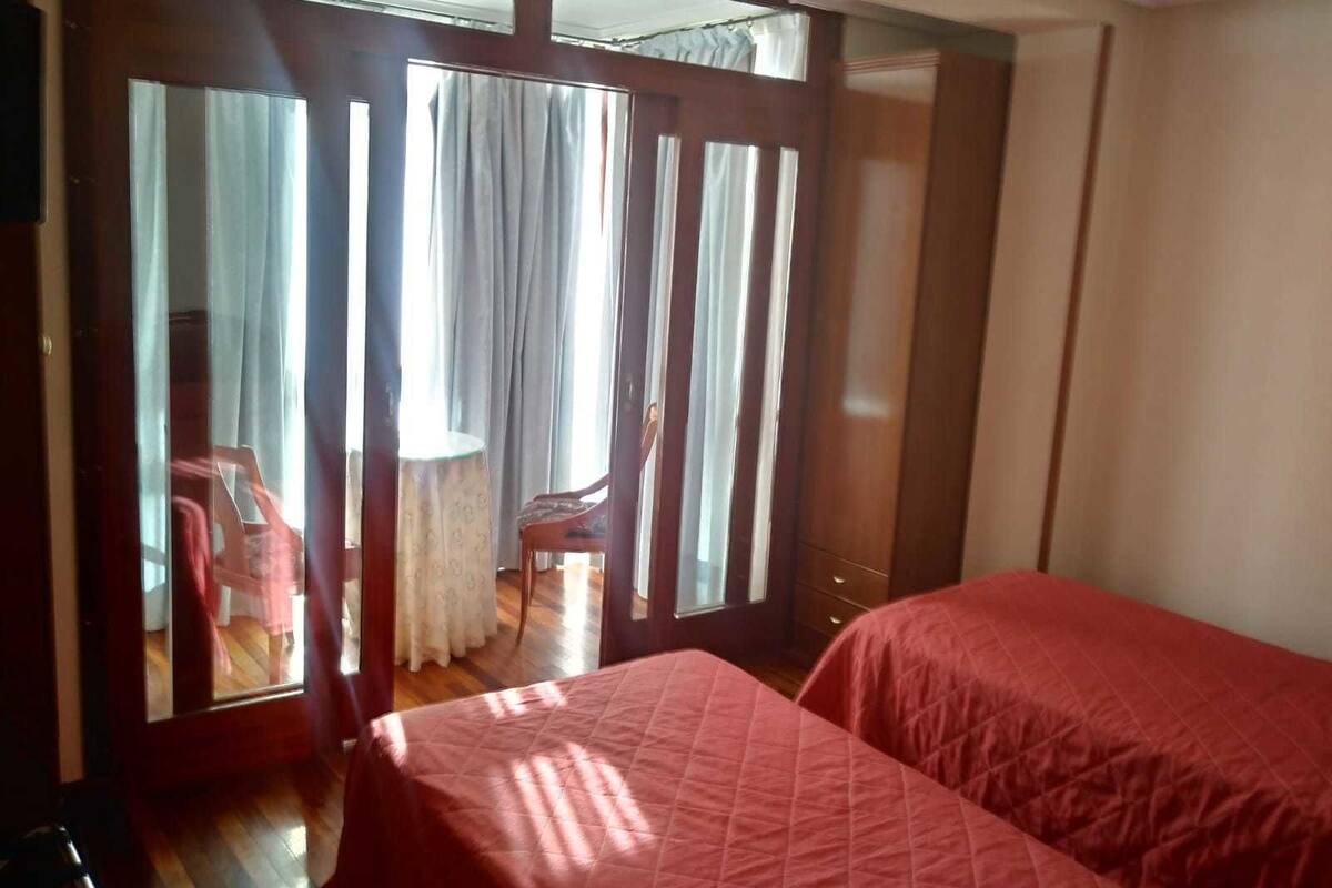 Una habitación con dos camas, un armario de madera, una mesita con una silla y una ventana con cortinas.