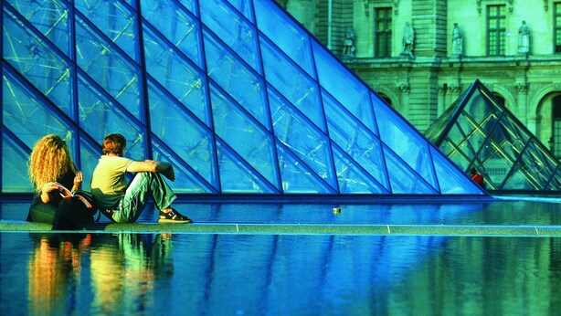 Two people sitting on the ground near a large glass structure with a reflection of a building in the background. - Le Carmin (Vitry-sur-Seine)