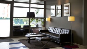 A modern lobby with a leather sofa, a coffee table, and a floor lamp. There are large windows and framed pictures on the wall. - Le Carmin (Vitry-sur-Seine)