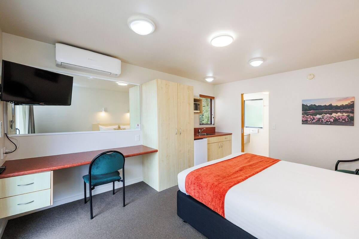 Quarto de hotel com cama, escrivaninha, cadeira, cozinha compacta e televisão.