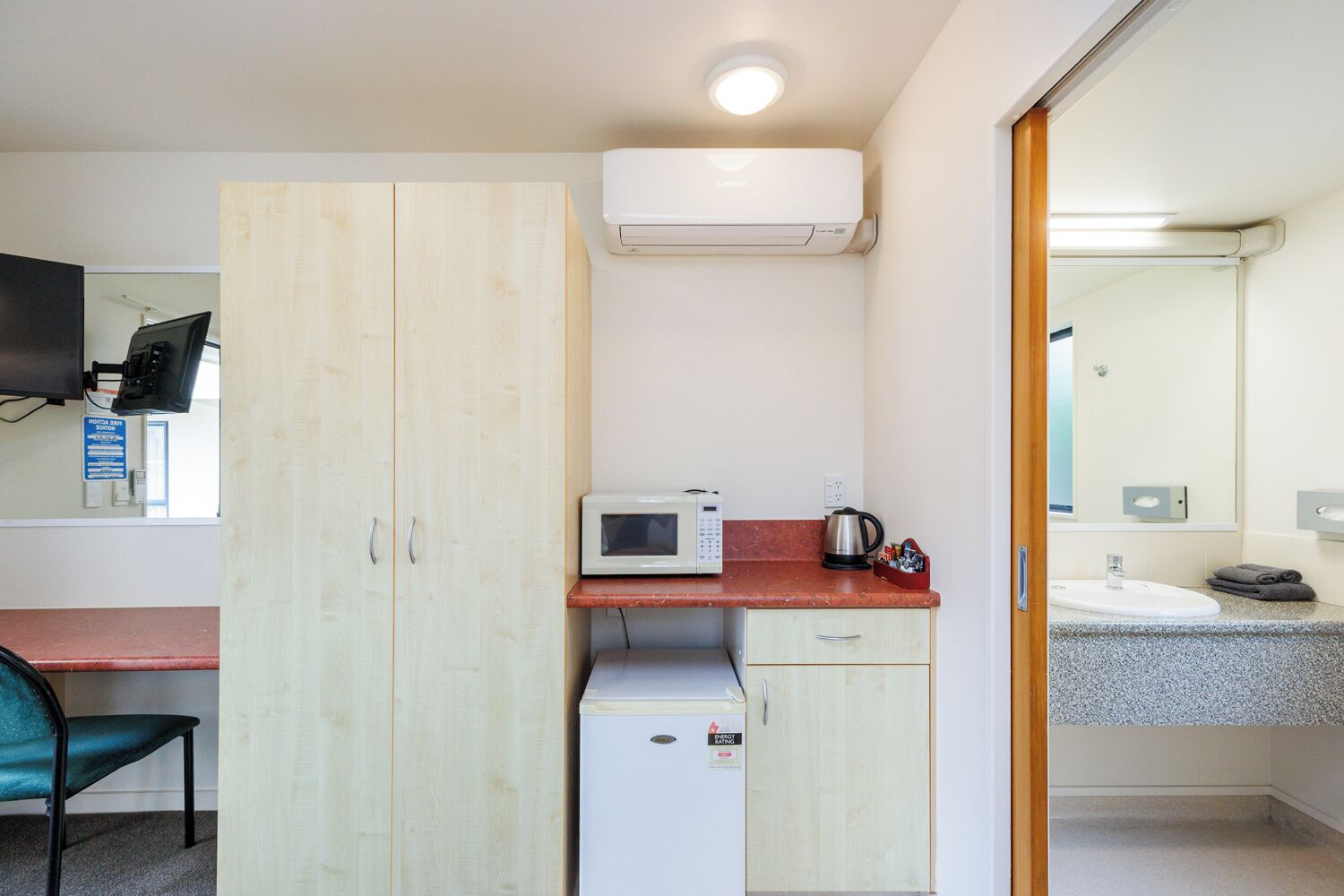 Una habitación compacta con kitchenette, microondas, refrigerador pequeño, armario de madera, escritorio con silla y baño con lavamanos y espejo.