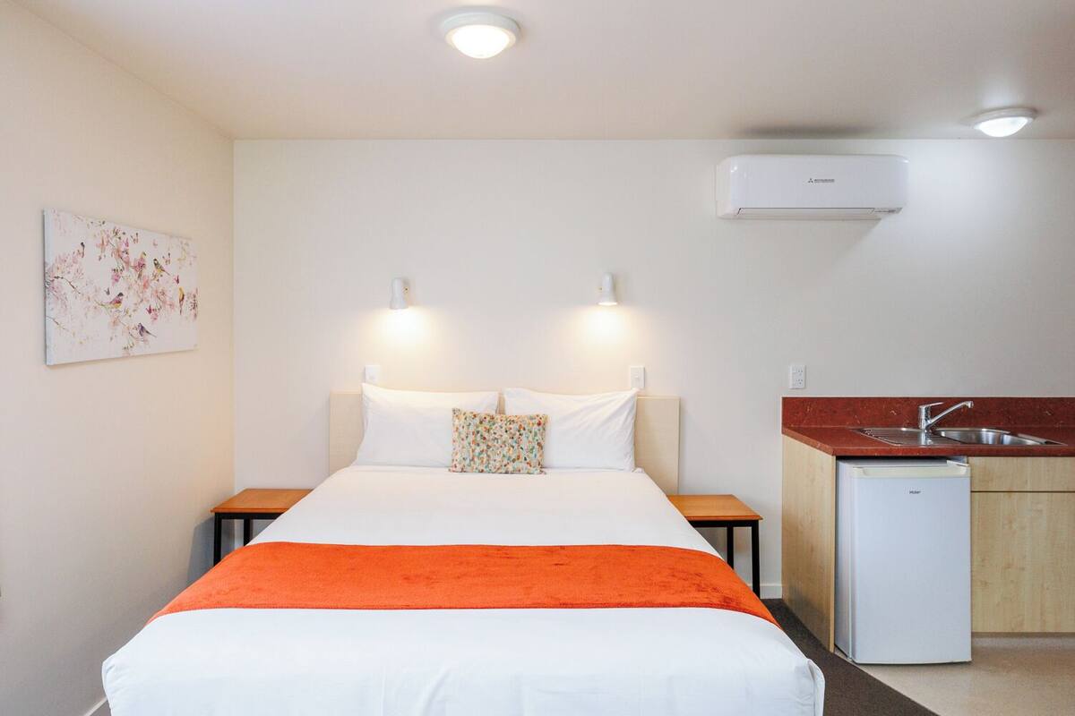 Quarto de hotel com uma cama, duas mesas de cabeceira, um aparador, uma pequena cozinha com pia e geladeira, e um ar condicionado na parede.
