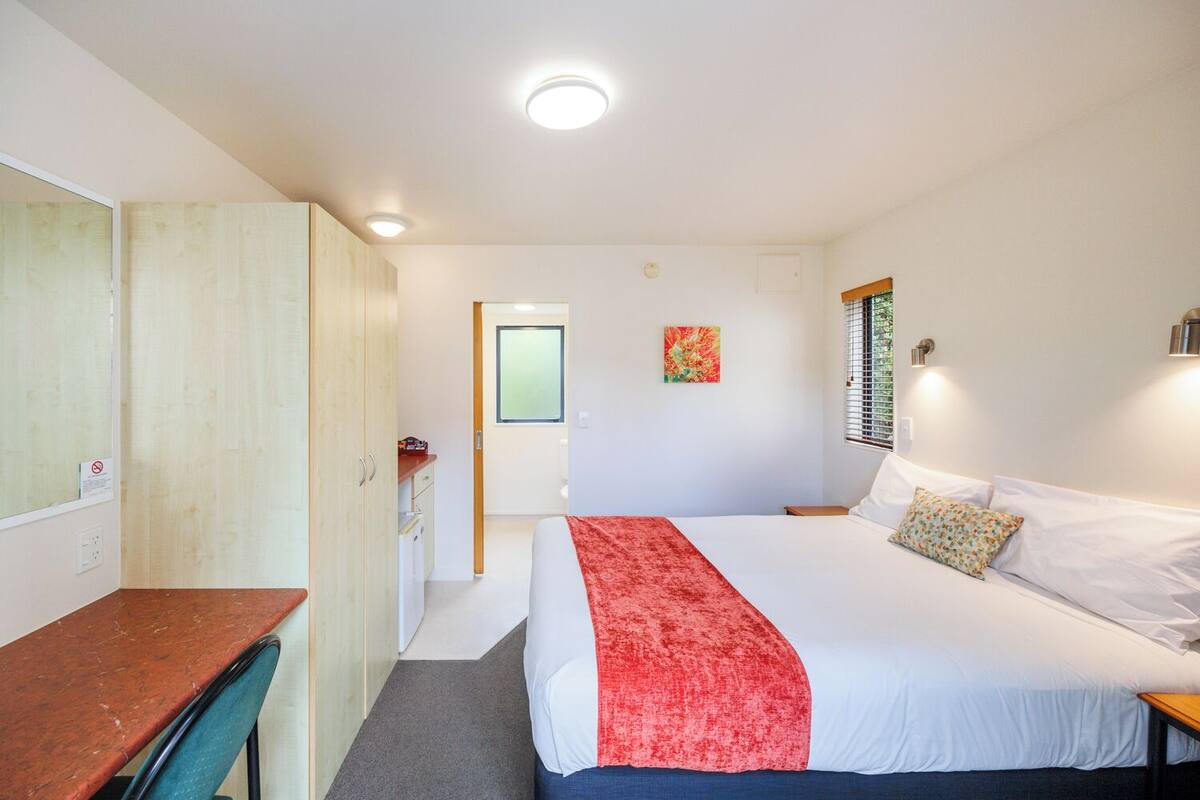 Quarto com cama, escrivaninha, guarda-roupa, janela com persianas e porta que dá para outro cômodo.