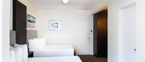 A hotel room with two single beds, a small desk, a chair, a wall-mounted TV, and a door leading to another room.
