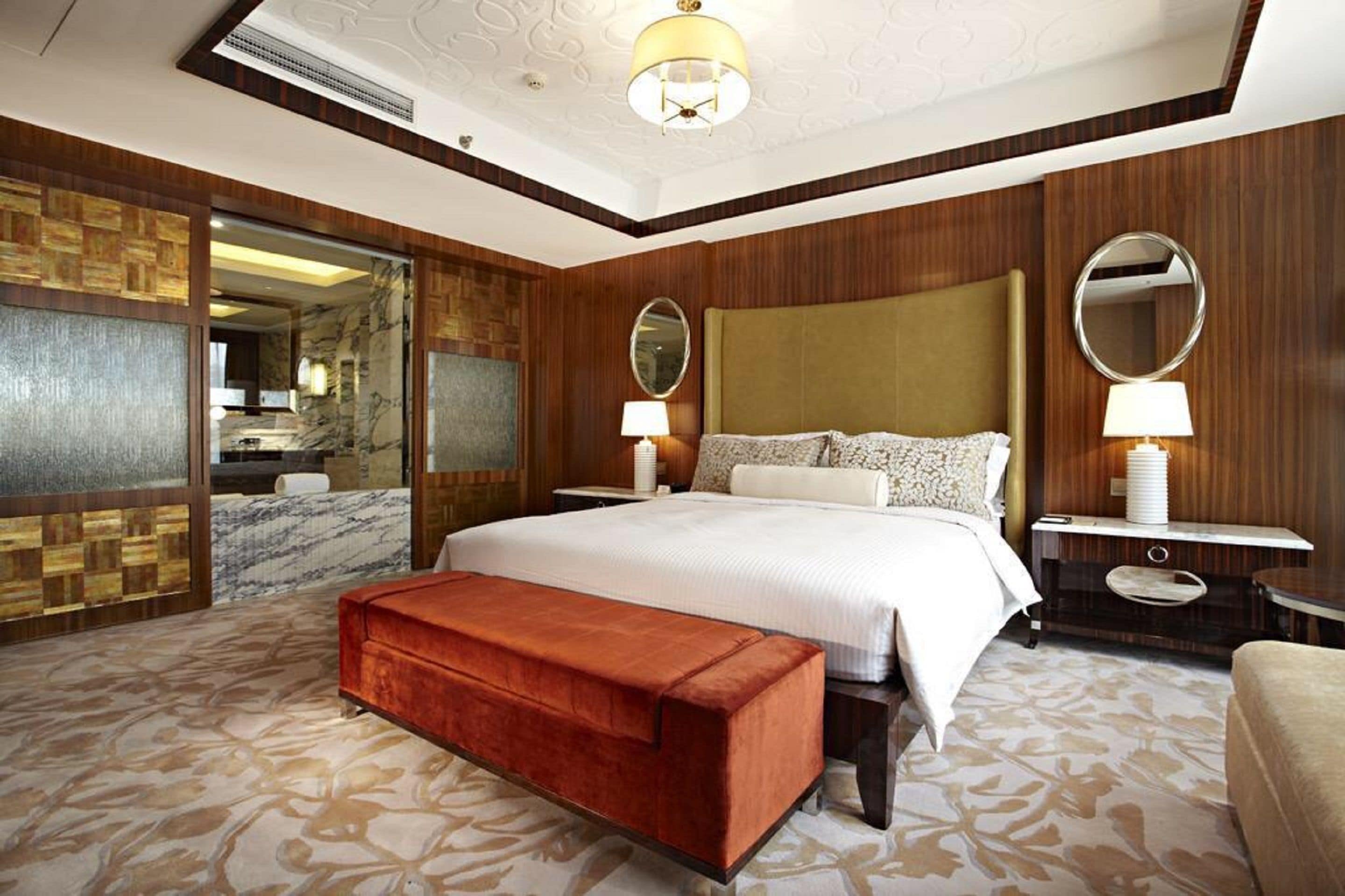 Habitación de hotel con una cama grande, dos mesitas de noche con lámparas, un cabecero de madera, un banco y un espejo amplio.