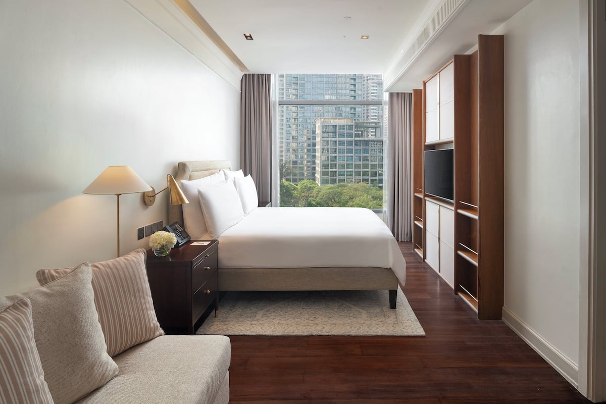 A modern hotel room with a large bed, a bedside table with a lamp, a sofa, and a view of a cityscape through floor-to-ceiling windows.