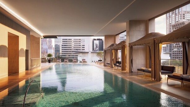 Una piscina interior con agua cristalina, rodeada de sillones de descanso y vistas a edificios altos.
