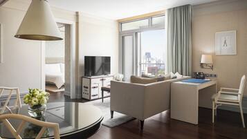 A modern hotel room with a sofa, a desk, a TV, and a large window with a city view.
