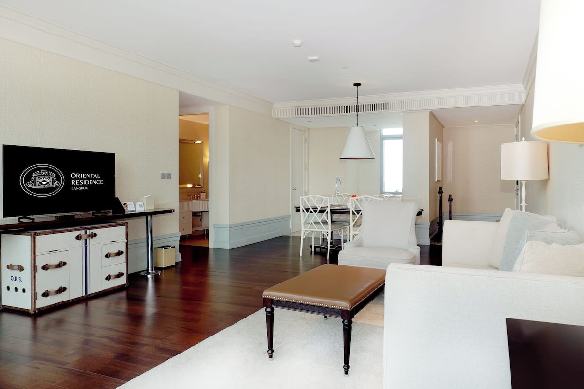 A modern living room with a flat-screen TV, a white sofa, a brown bench, and a dining area with a white table and chairs.