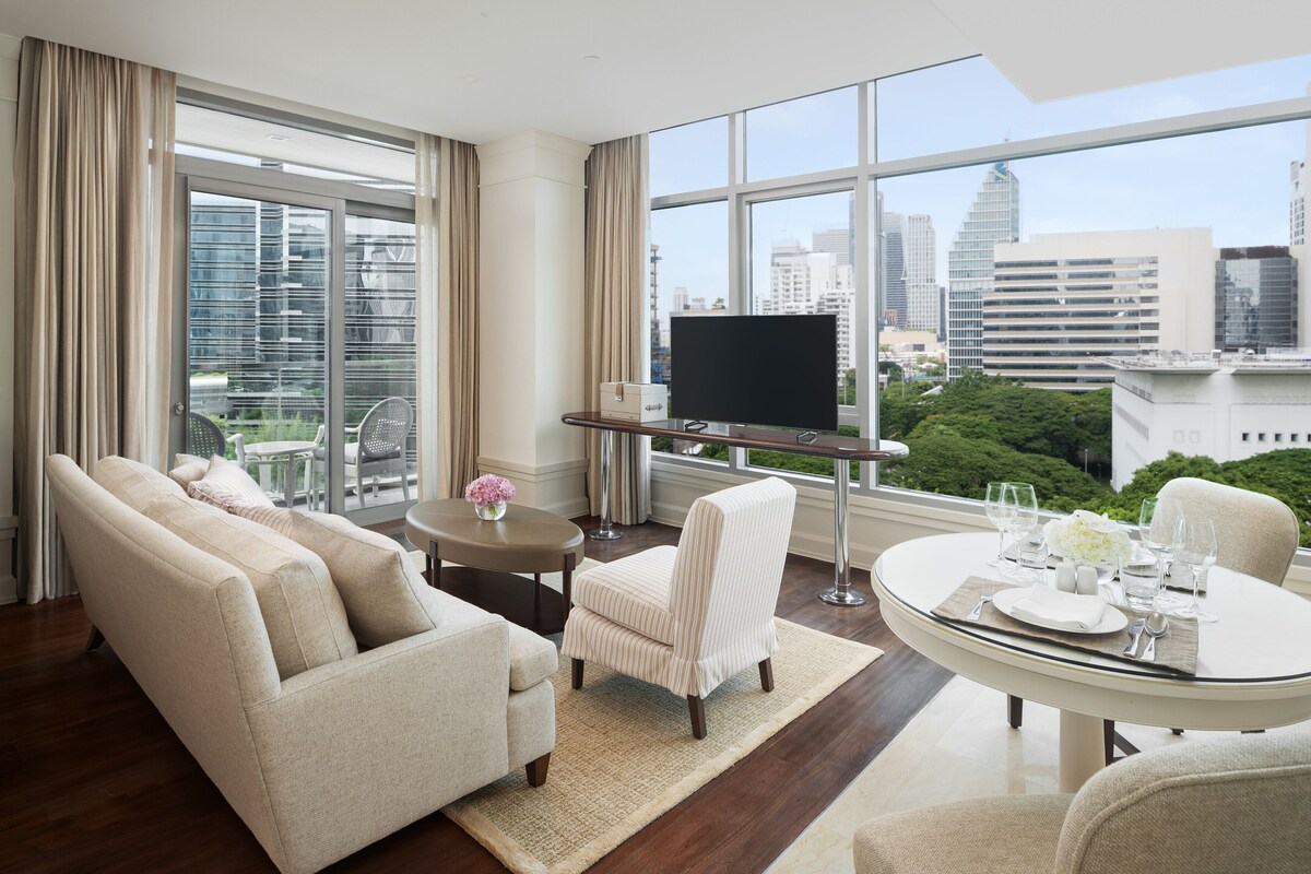 Una habitación de hotel moderna con sofá, sillones, una mesa de comedor para dos personas, televisor sobre un mueble y grandes ventanales con vista a la ciudad.