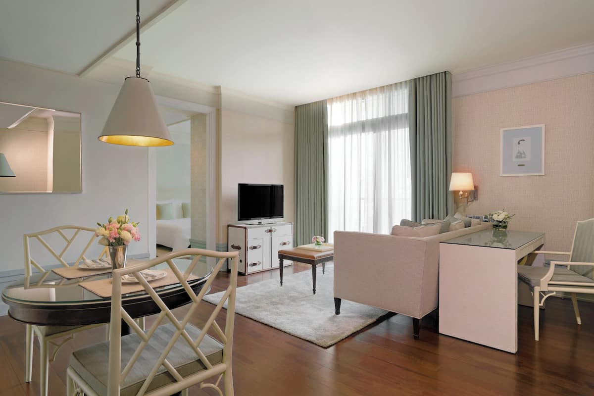 A modern hotel room with a glass dining table, a sofa, a TV, and a desk with a lamp.