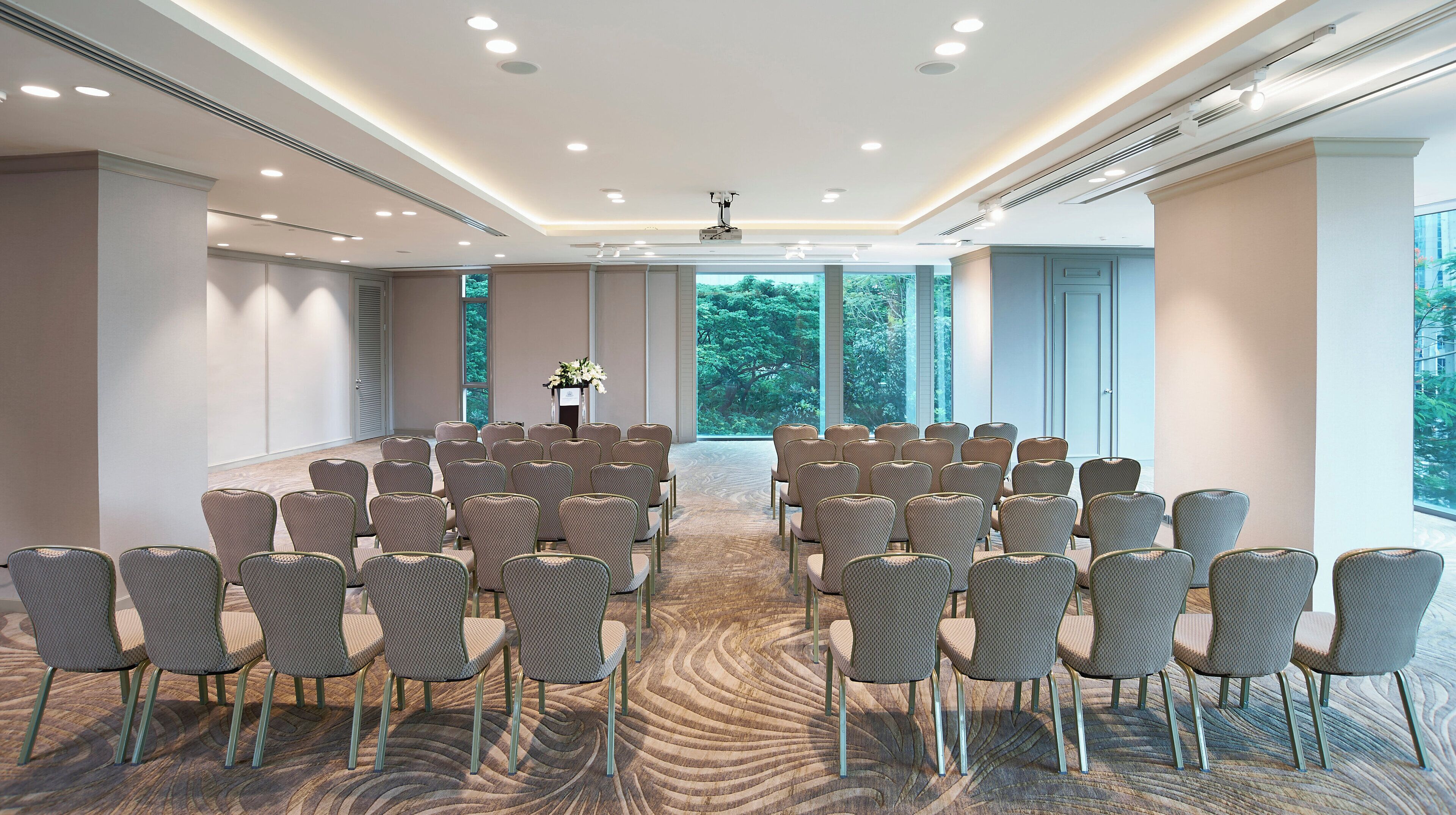 A conference room with rows of chairs facing a podium, large windows offering a view of trees, and a carpeted floor.