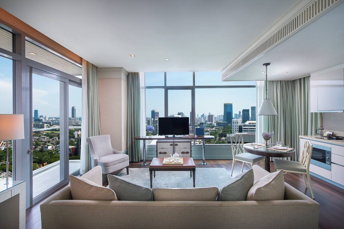 A modern hotel room with a large window offering a city view, a sofa, a dining table, and a kitchenette.