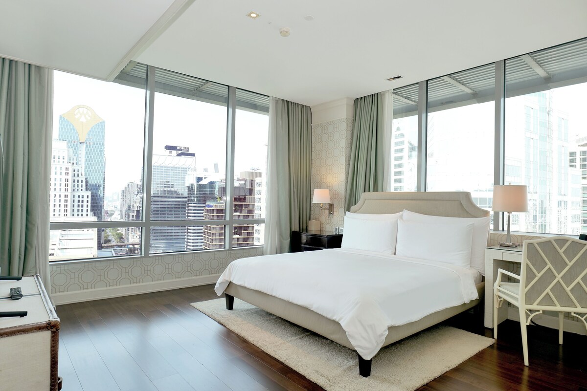 Une chambre d’hôtel moderne, dotée d’un grand lit, d’un bureau et d’une chaise. La chambre dispose de grandes fenêtres offrant une vue sur la ville.