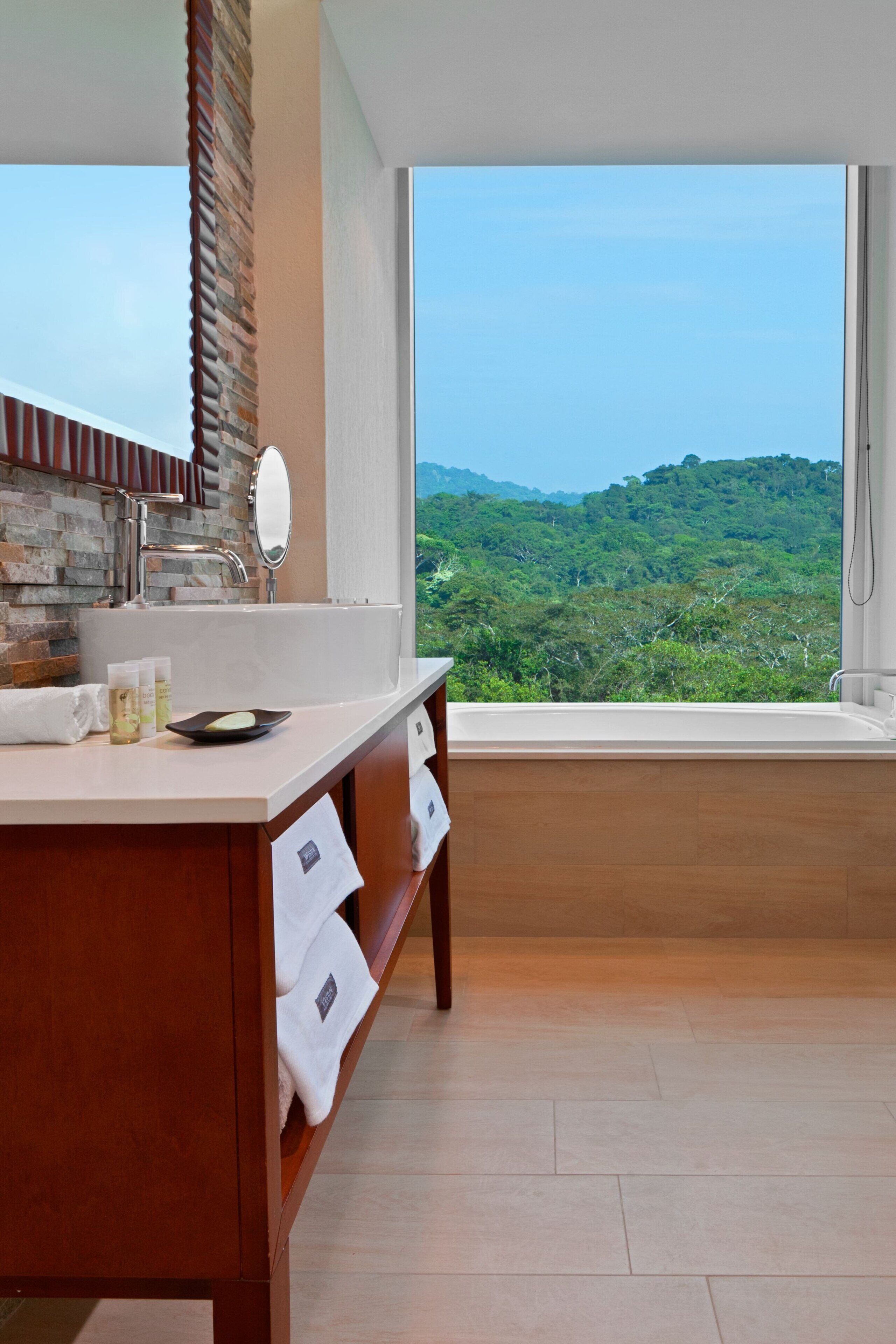 A bathroom with a large window, a wooden vanity, a mirror, and toiletries.