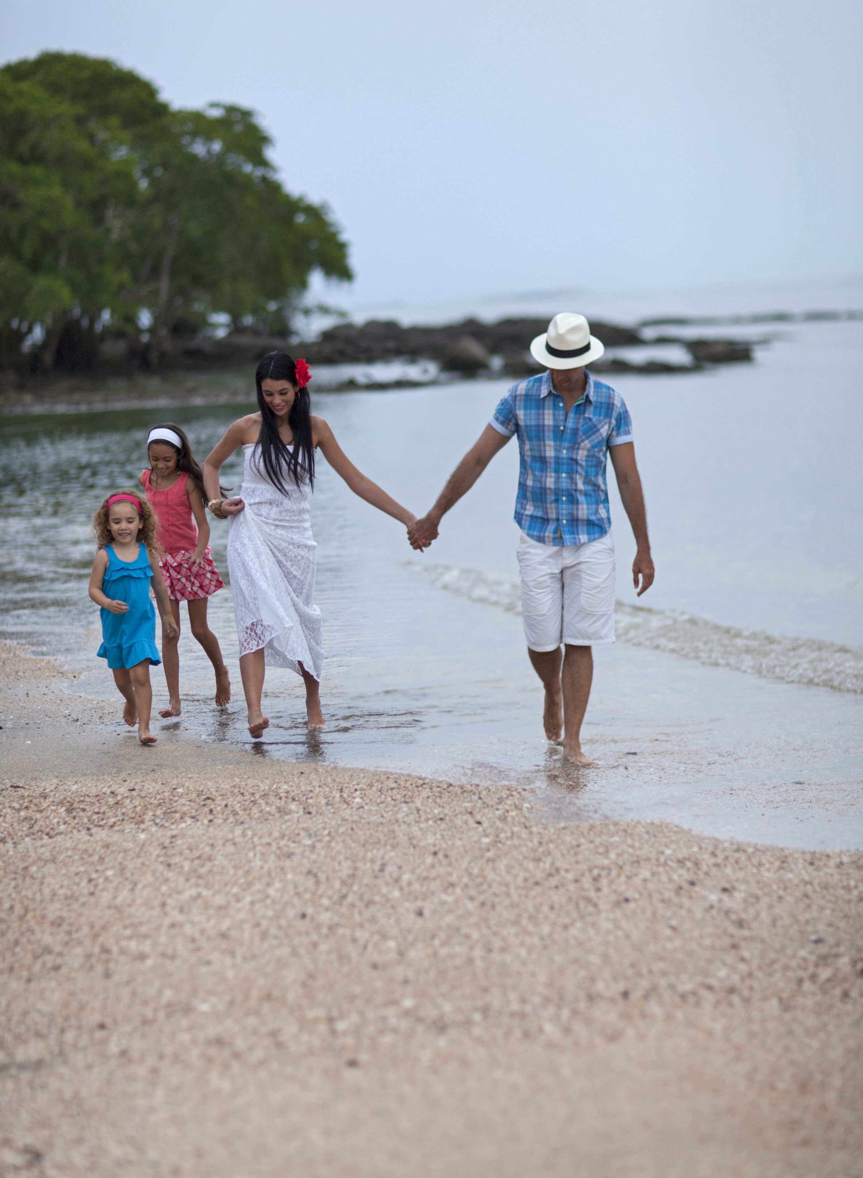 Una familia caminando sobre una playa de guijarros con agua tranquila y vegetación al fondo.