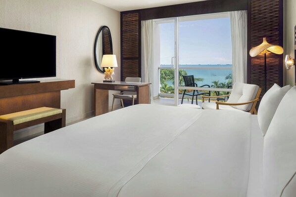 Habitación de hotel con una cama grande, un escritorio, una silla, un televisor, una lámpara y un balcón con vista al mar.