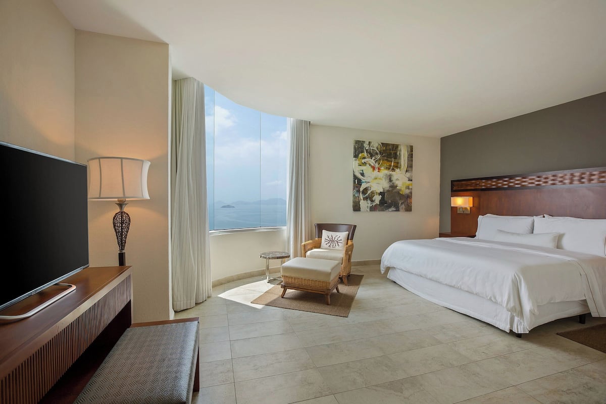 A hotel room with a large bed, a TV, a chair, and a view of the ocean.