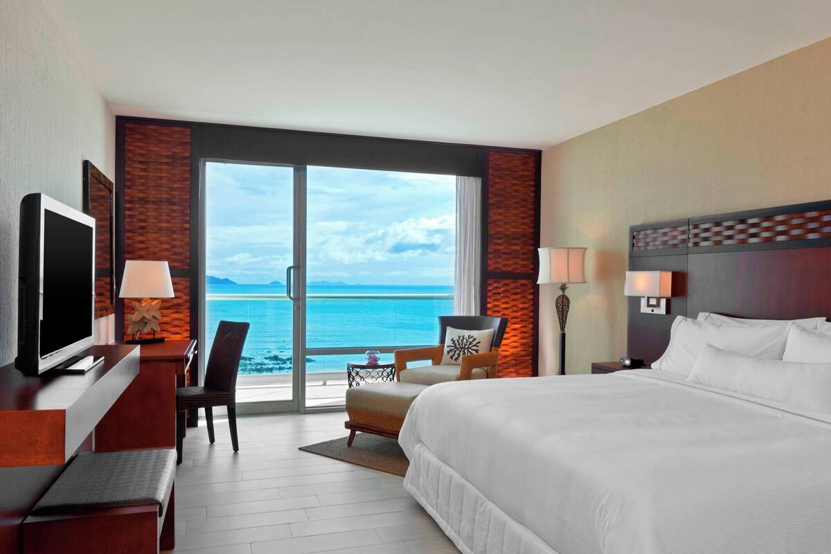 A hotel room with a large bed, a desk with a TV, a chair, and a view of the ocean through a sliding door.