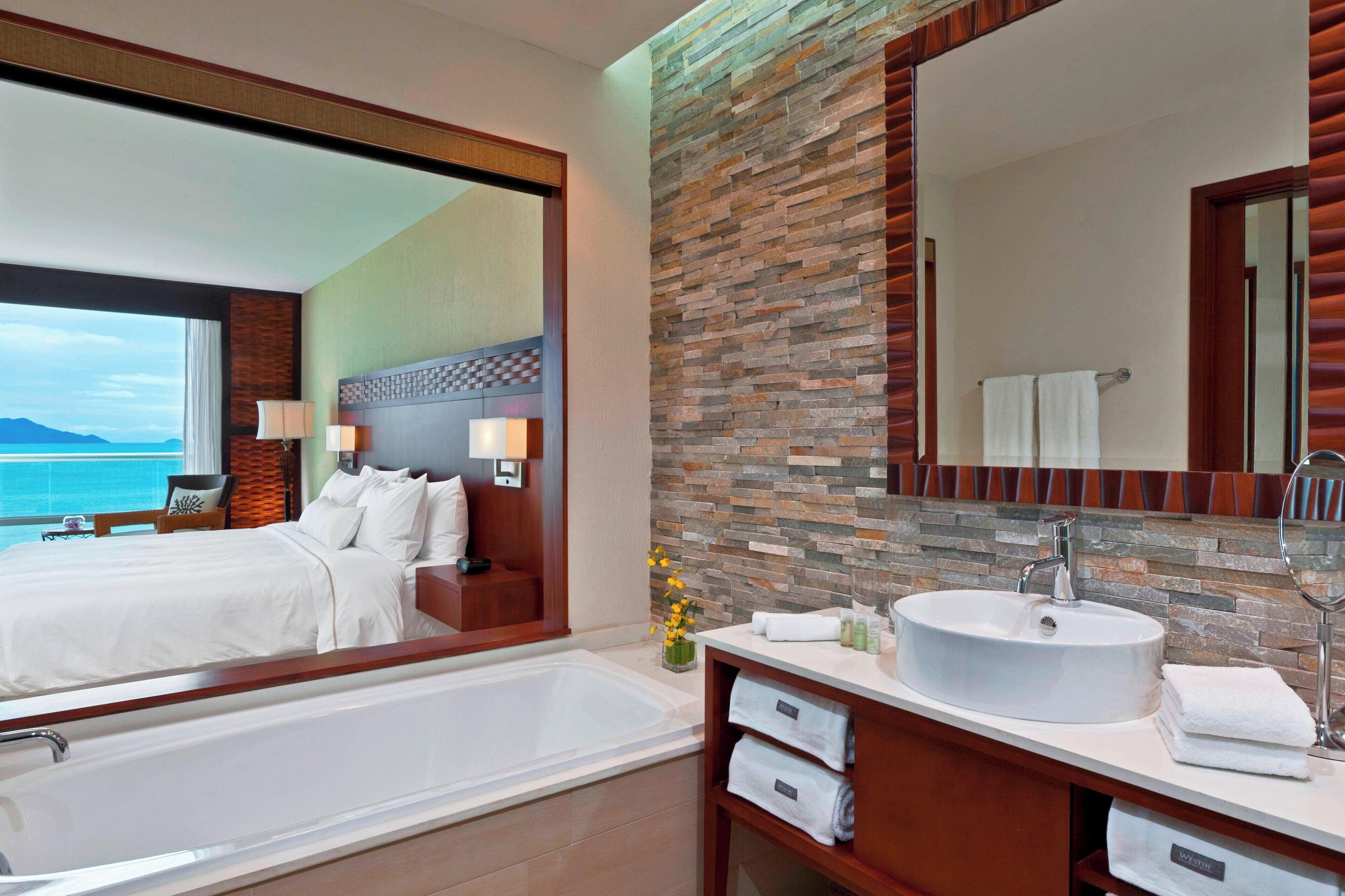 A modern hotel bathroom with a large bathtub, a round mirror, and a stone wall backsplash.