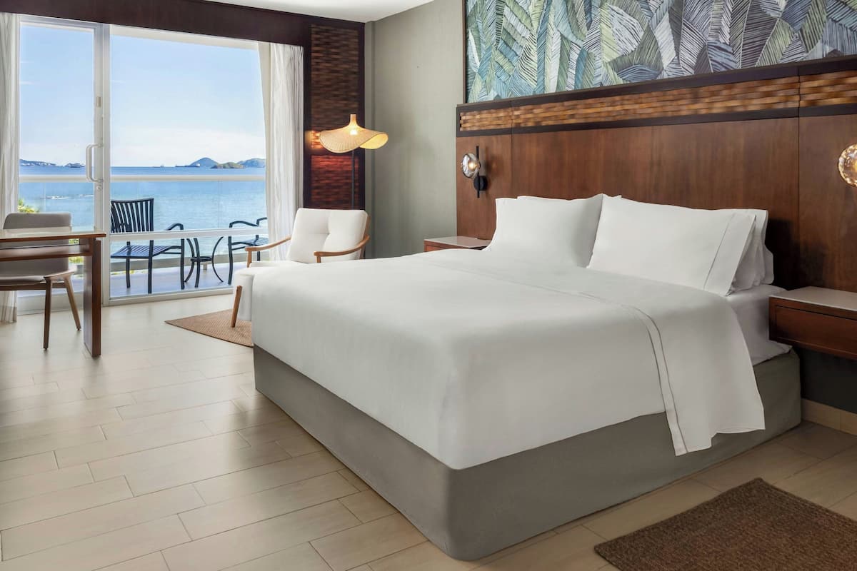 A hotel room with a large bed, a desk, a chair, and a view of the sea.